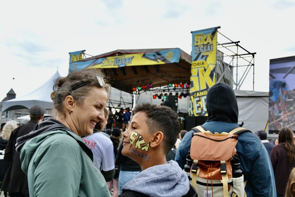 A mom and her son stand back from the stage at the Ska and Reggae Festival in Victoria. The boys face paint on his cheek matches the stage sign and says Ska and Reggae. Photography by Rhonda Jesckey