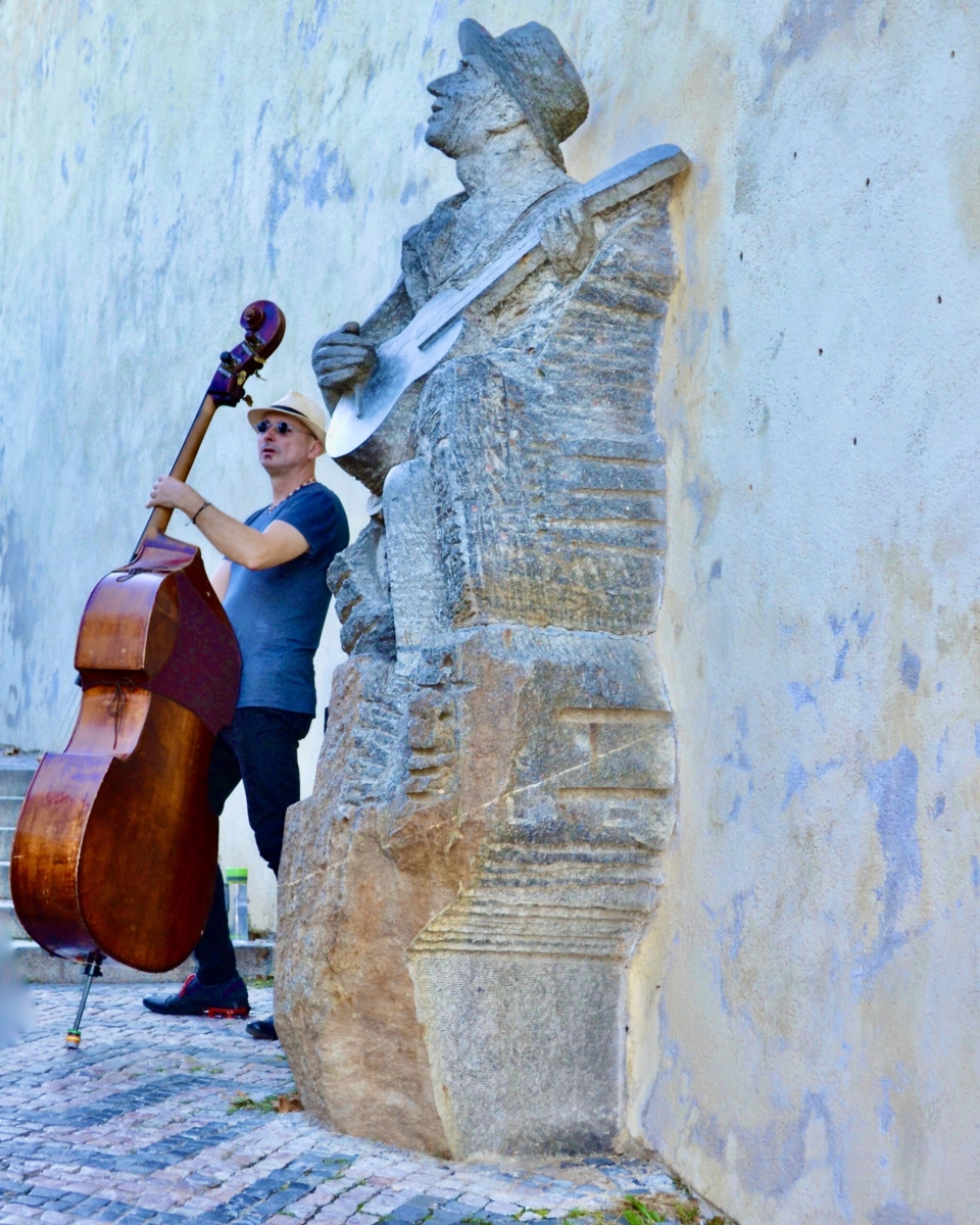 Man playing a bass standing beside a stone sculpture of a man playing a guitar in Prague. Photography by Rhonda Jesckey