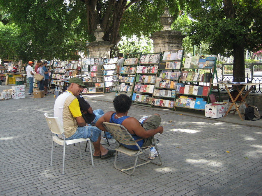 Three booksellers sitting in chairs and chatting in front of book racks where 5 people in the distance browse. Photography by Rhonda Jesckey