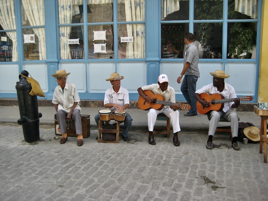 Four older musicians sit and play music on the street in Havana - 2 guitars and a bongo player and one without an instrument. Photography by Rhonda Jesckey